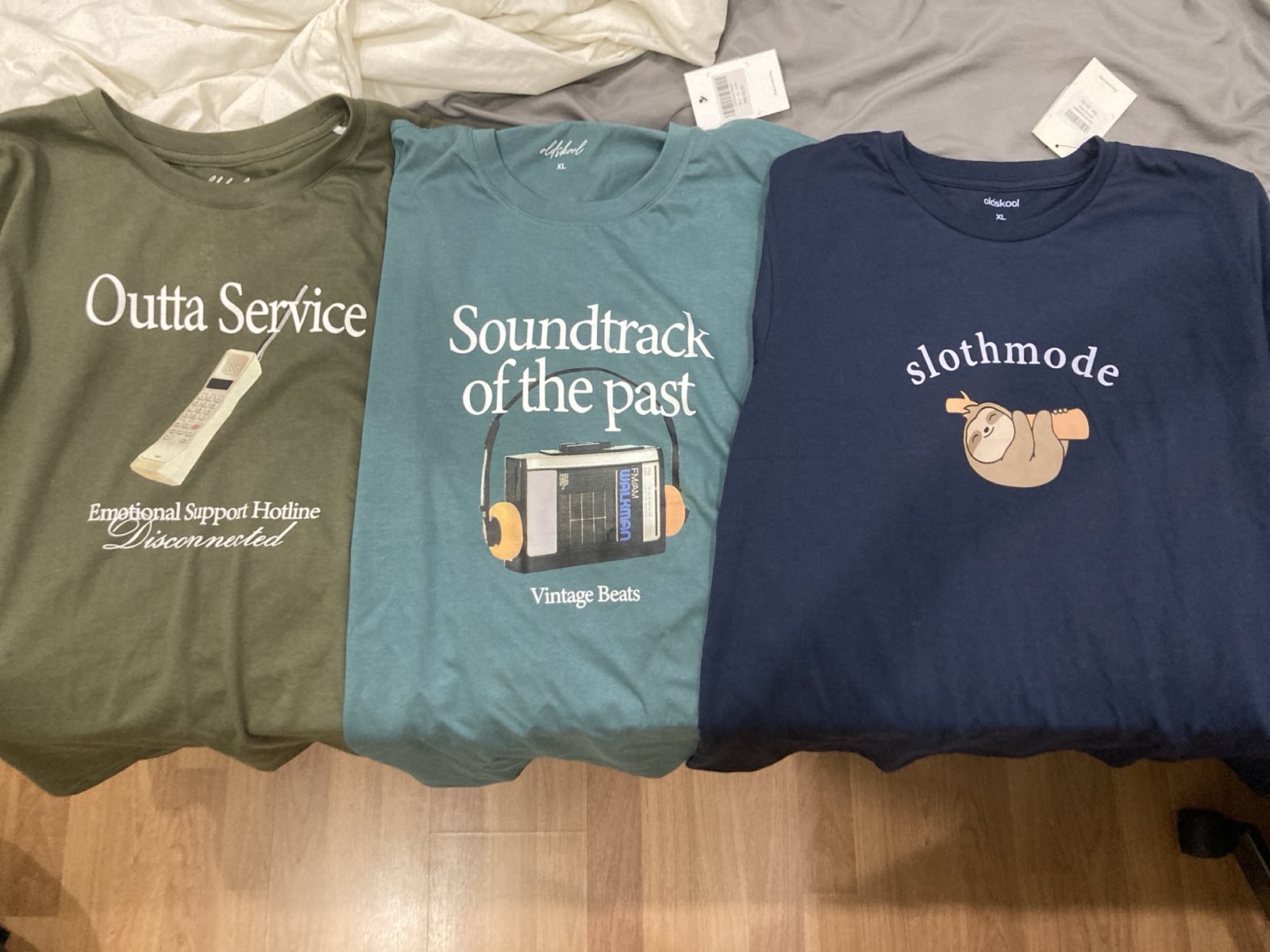 Three tee shirts, side by side. The first reads "Outta Service: Emotional Support Hotline Disconnected" and has a picture of an old cell phone. The second reads "Soundtrack of the past: Vintage Beats" with a picture of a Walkman. The third shows a sloth hanging from a branch and the word "slothmode".